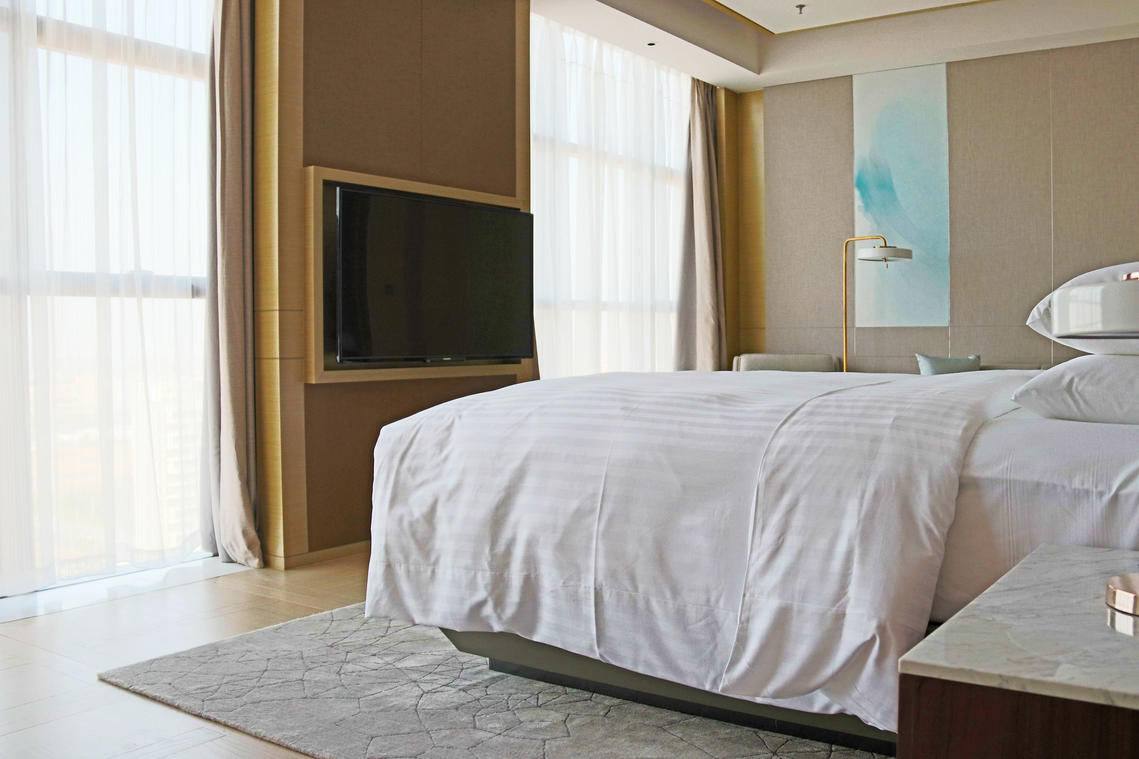 Een moderne hotelkamer met een groot bed, een flatscreen televisie aan de muur en een groot raam met dunne gordijnen.
