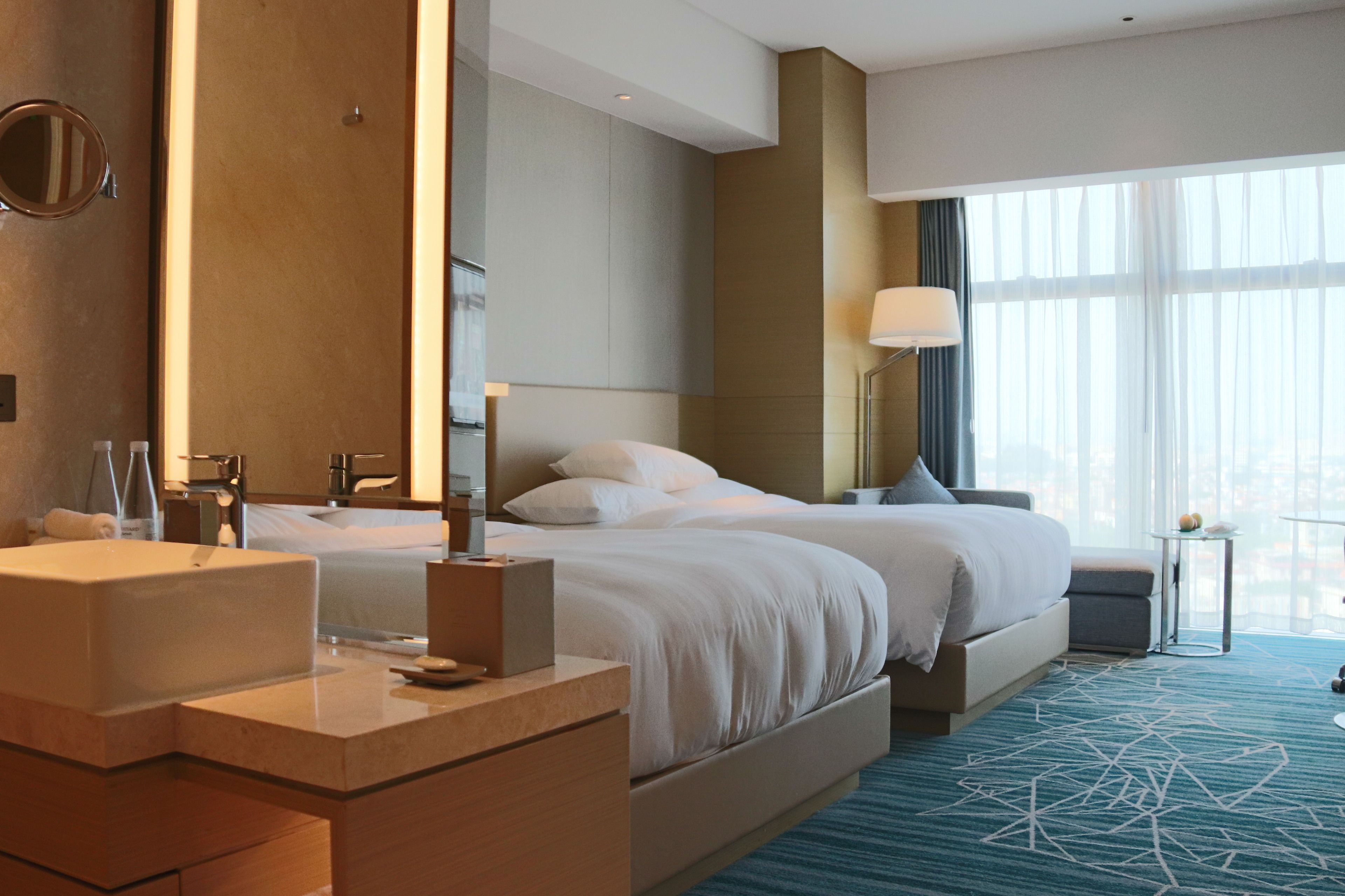 A hotel room with a large bed, a separate seating area, a bathroom with a sink and mirror, and a view of the city through large windows.