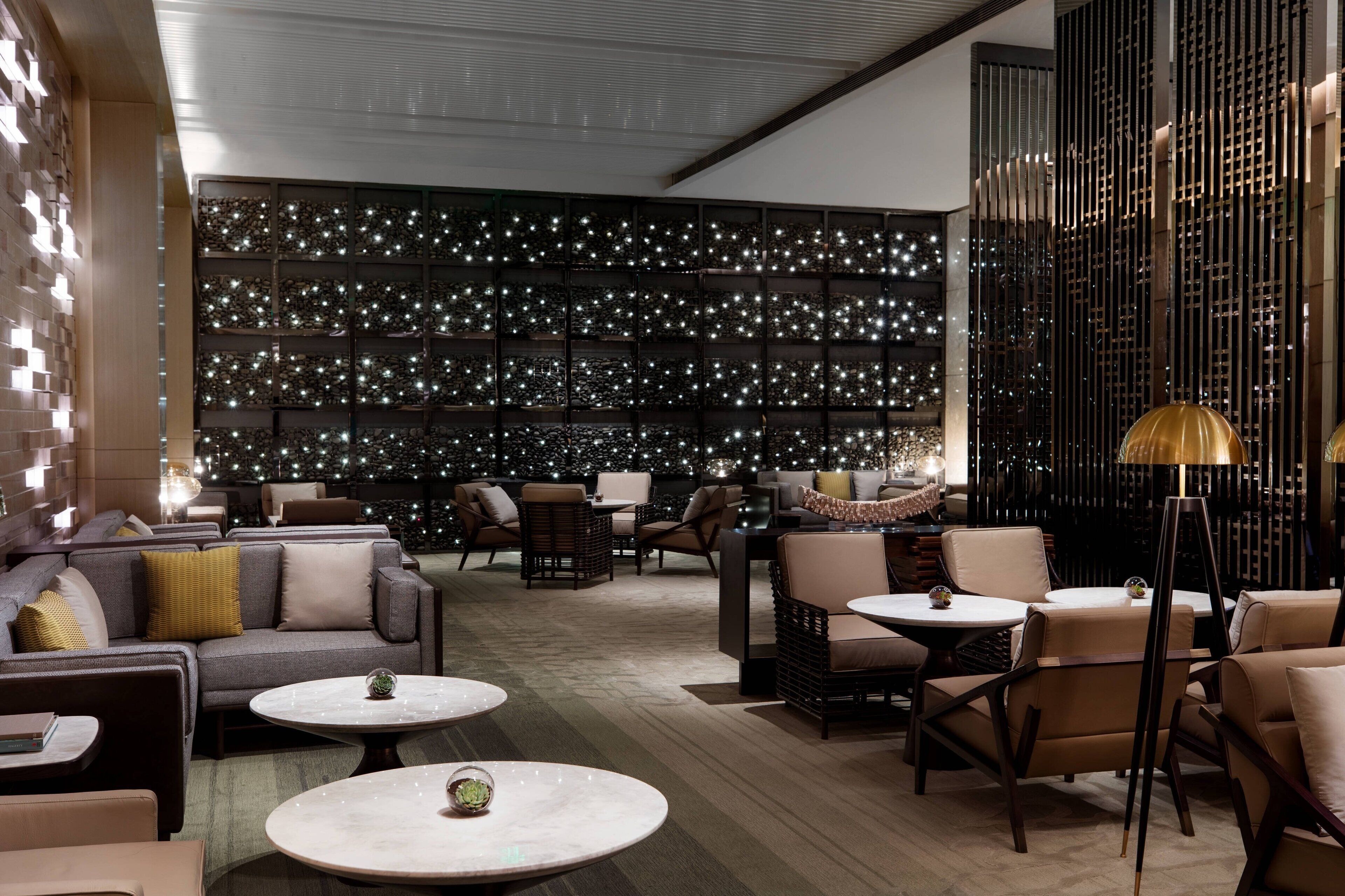 Ein stilvoller Lounge-Bereich mit Sofas, Sesseln und Tischen, der durch eine dekorative Wand mit Beleuchtung und eine hohe, schlanke Säulenstruktur ergänzt wird.