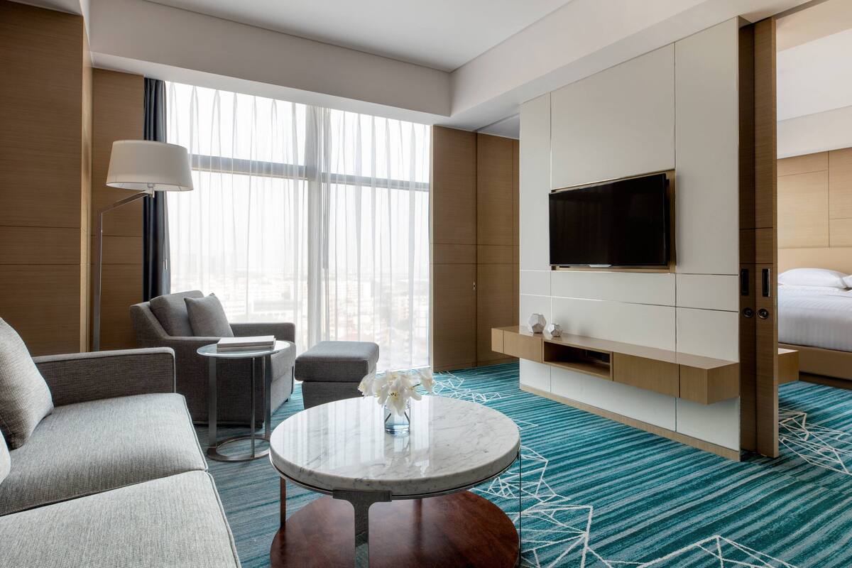 Quarto de hotel moderno com sofá, poltrona, mesa de centro redonda e televisão fixada num painel de madeira.