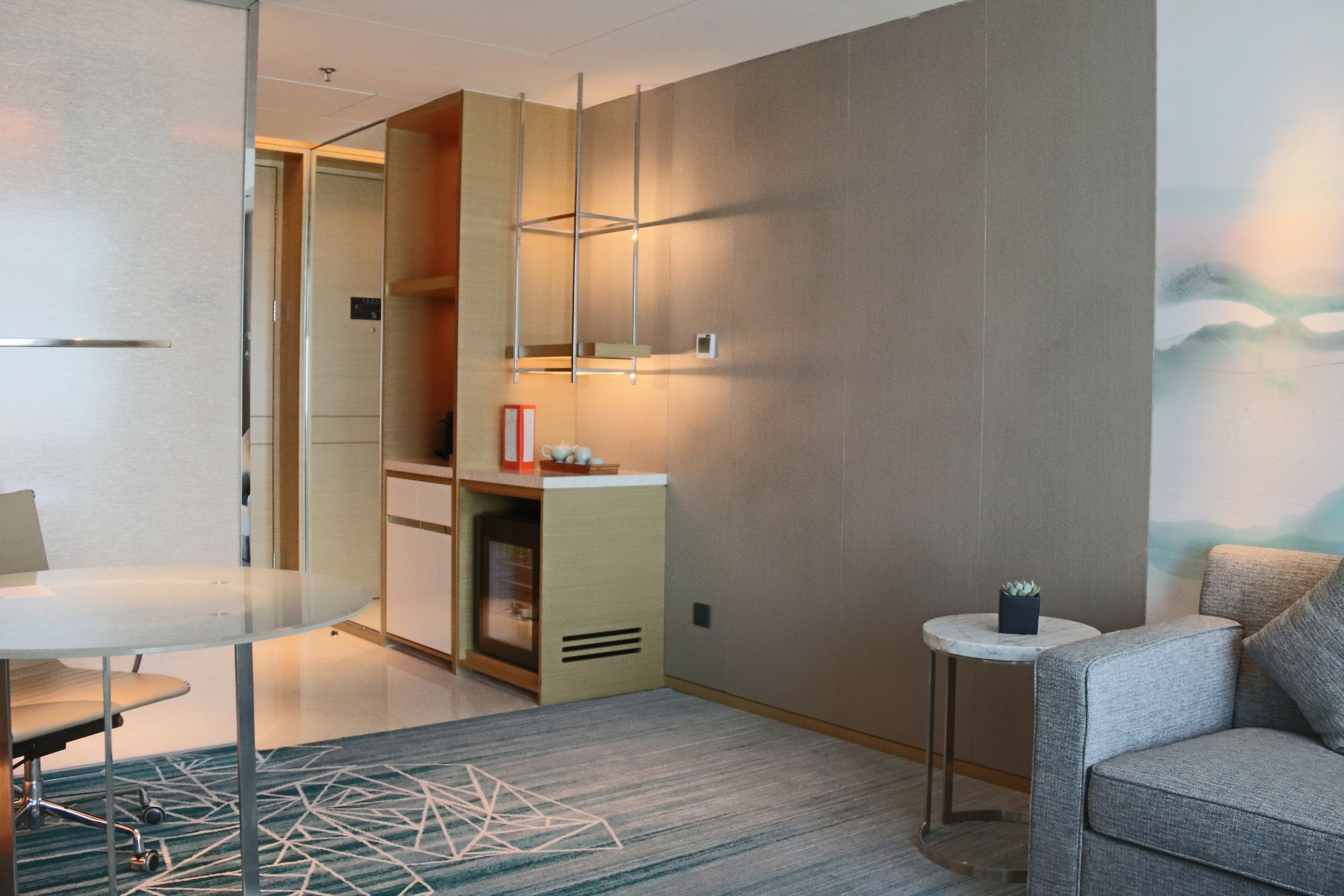 Een moderne hotelkamer met een glazen tafel, een bank, een klein bijzettafeltje met een plant en een wandplank met decoratieve objecten.
