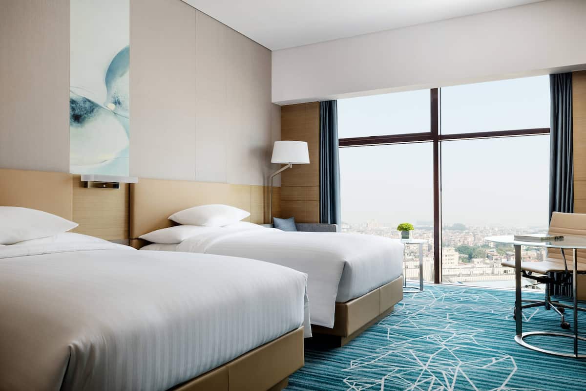 Quarto de hotel com duas camas, uma escrivaninha, uma luminária e uma janela grande com vista para a cidade.