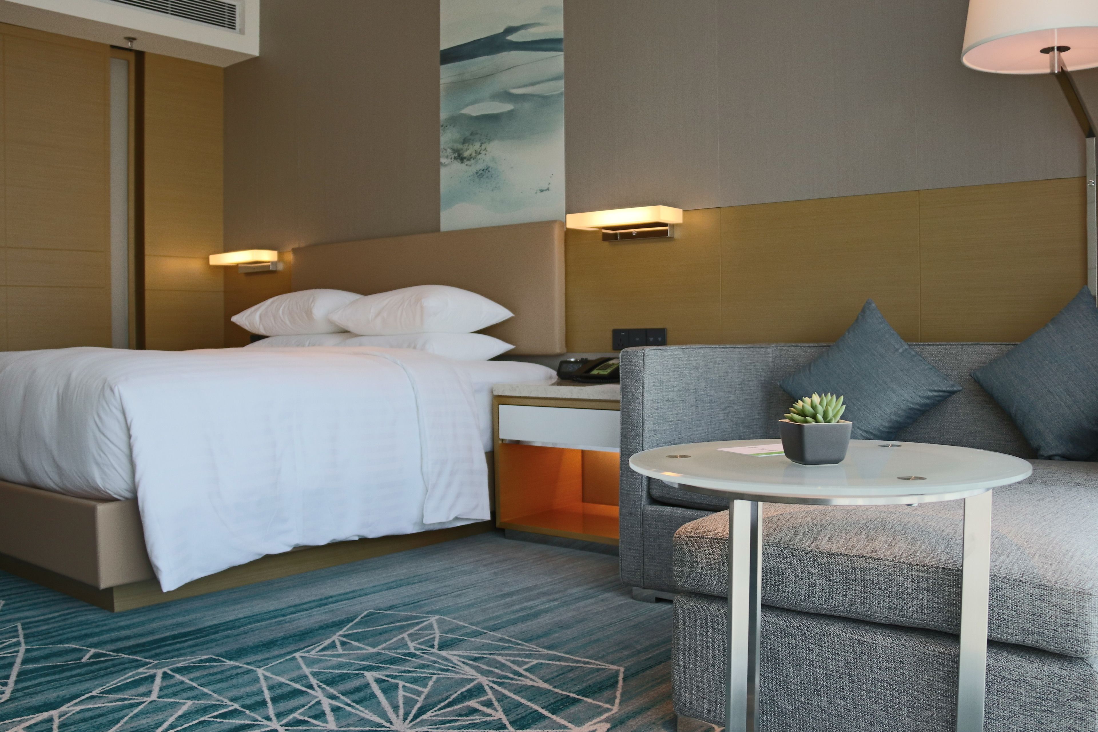 A hotel room with a bed, a sofa, a small table with a plant, and a wall-mounted lamp.