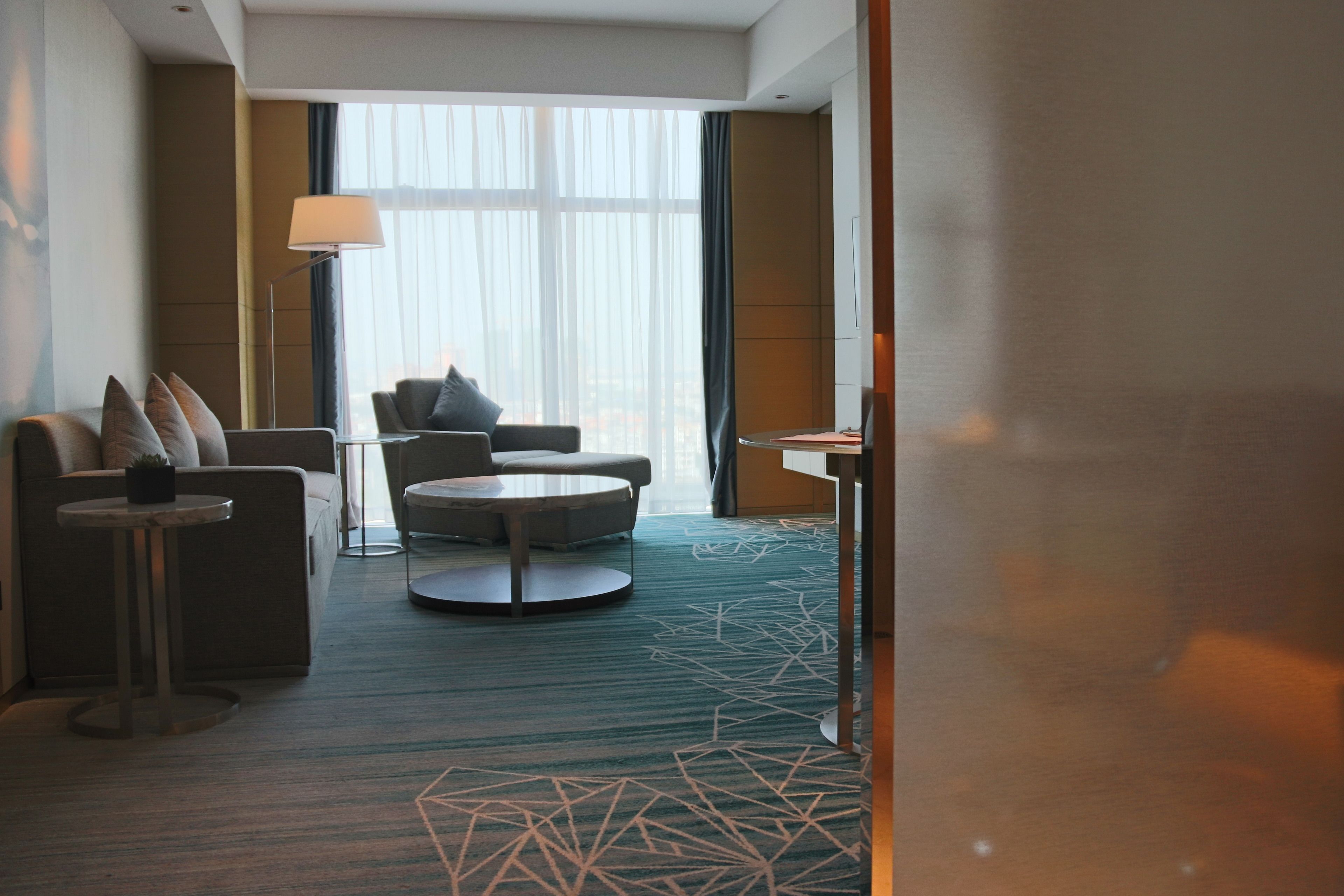 A modern hotel room with a sofa, armchair, round table, and a lamp.