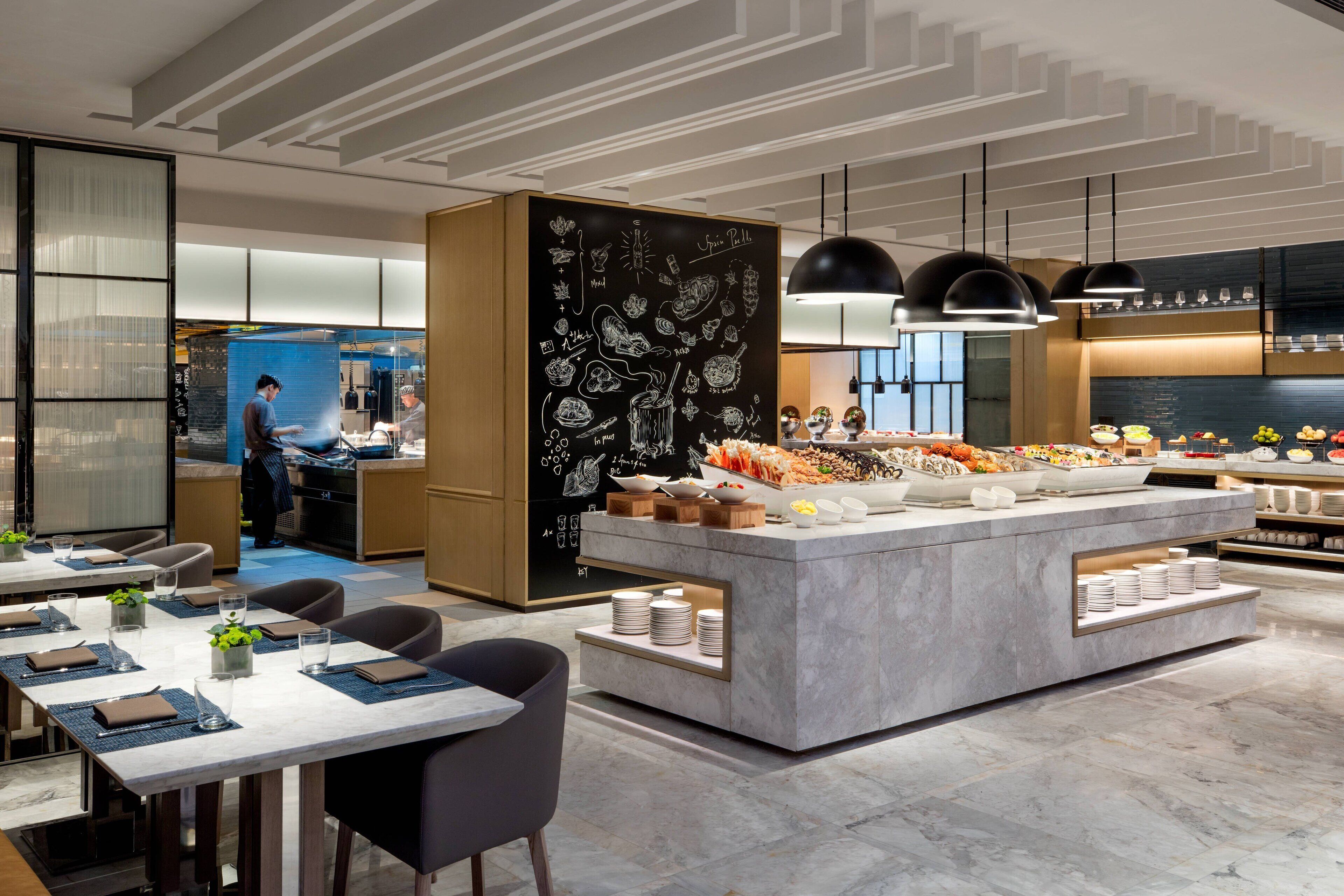 Un restaurante moderno con una zona de bufé central, mesas dispuestas y una cocina a la vista.