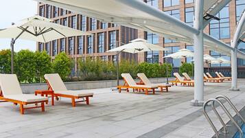 A rooftop pool area with sun loungers and umbrellas, a pool with a metal ladder, and a building with large windows in the background.