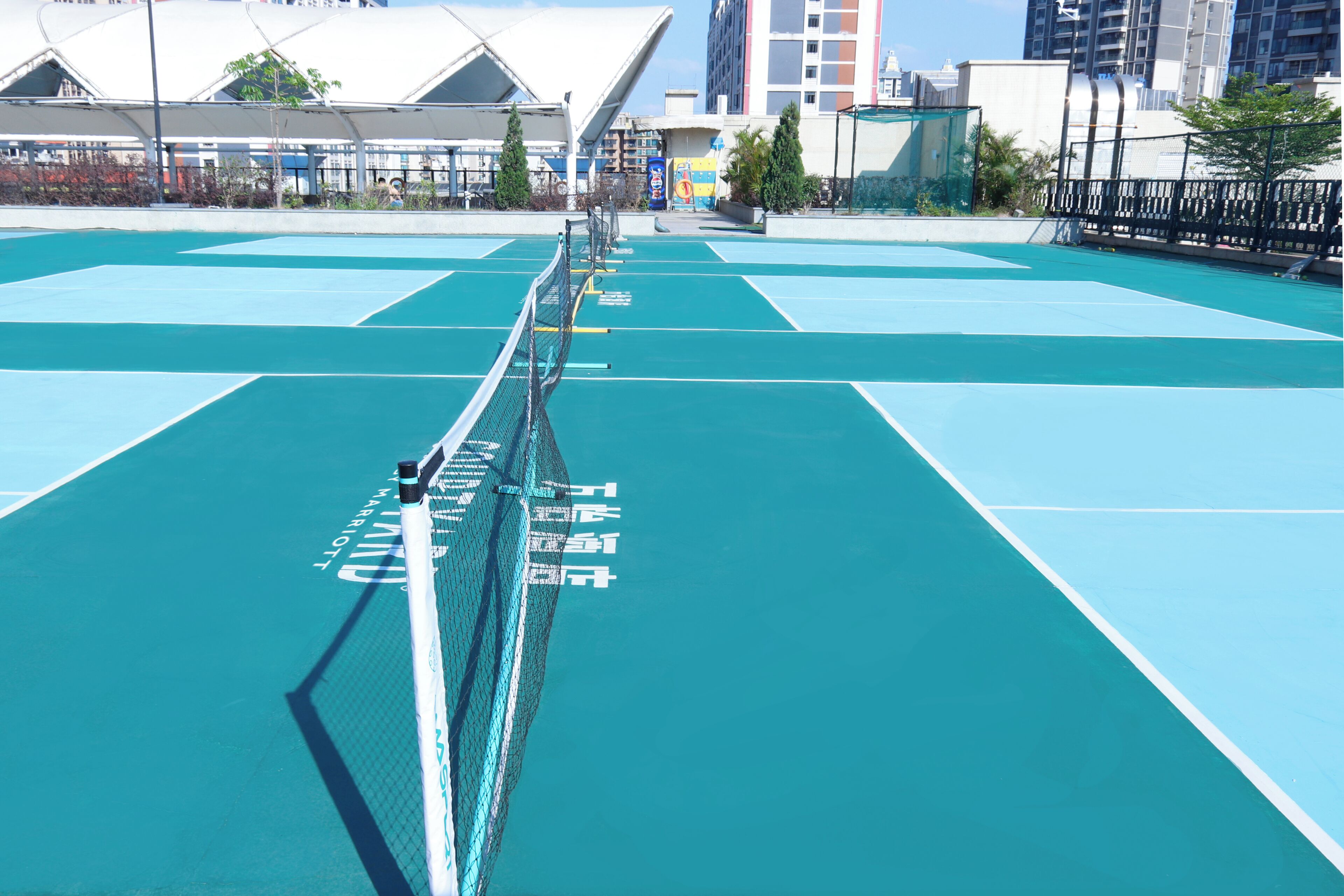 En tennisplan med ett nät i mitten, omgiven av ett staket och med byggnader i bakgrunden.