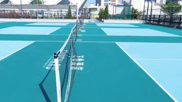 A tennis court with a net in the center, surrounded by a fence and buildings in the background.