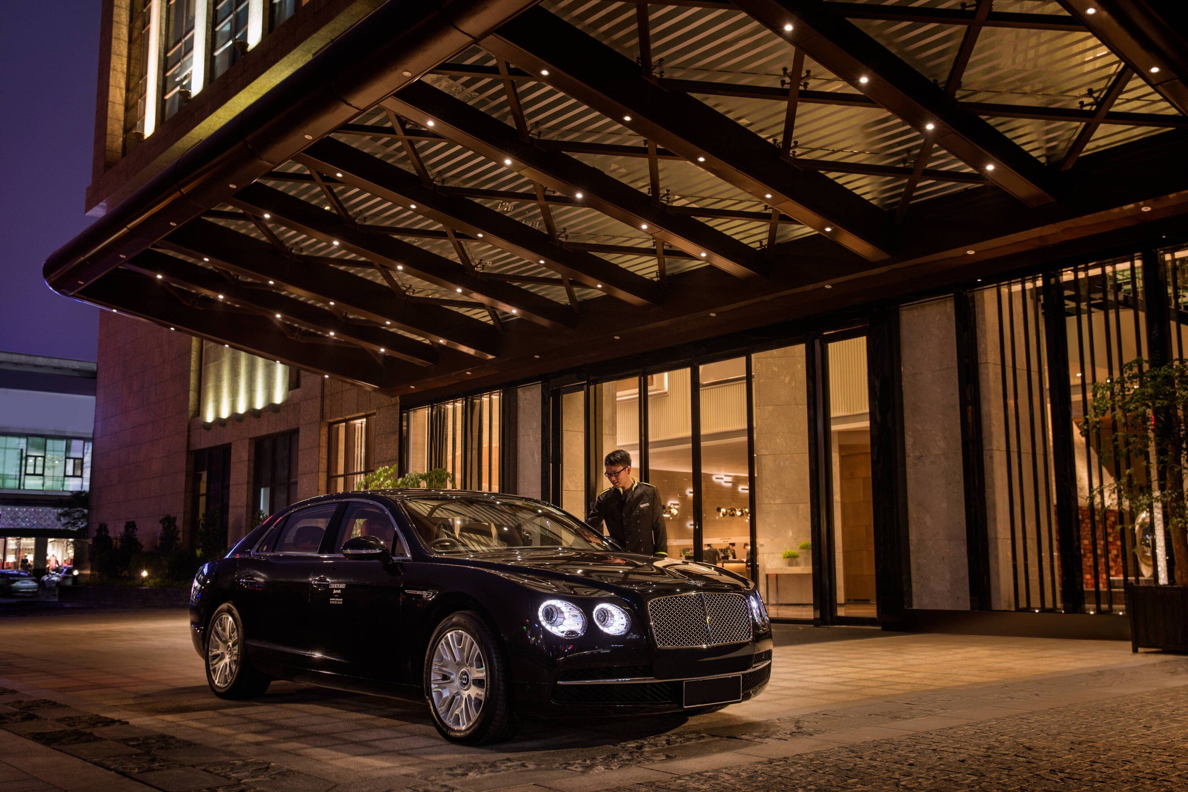 A black luxury car parked in front of a building with a covered entrance and large windows.