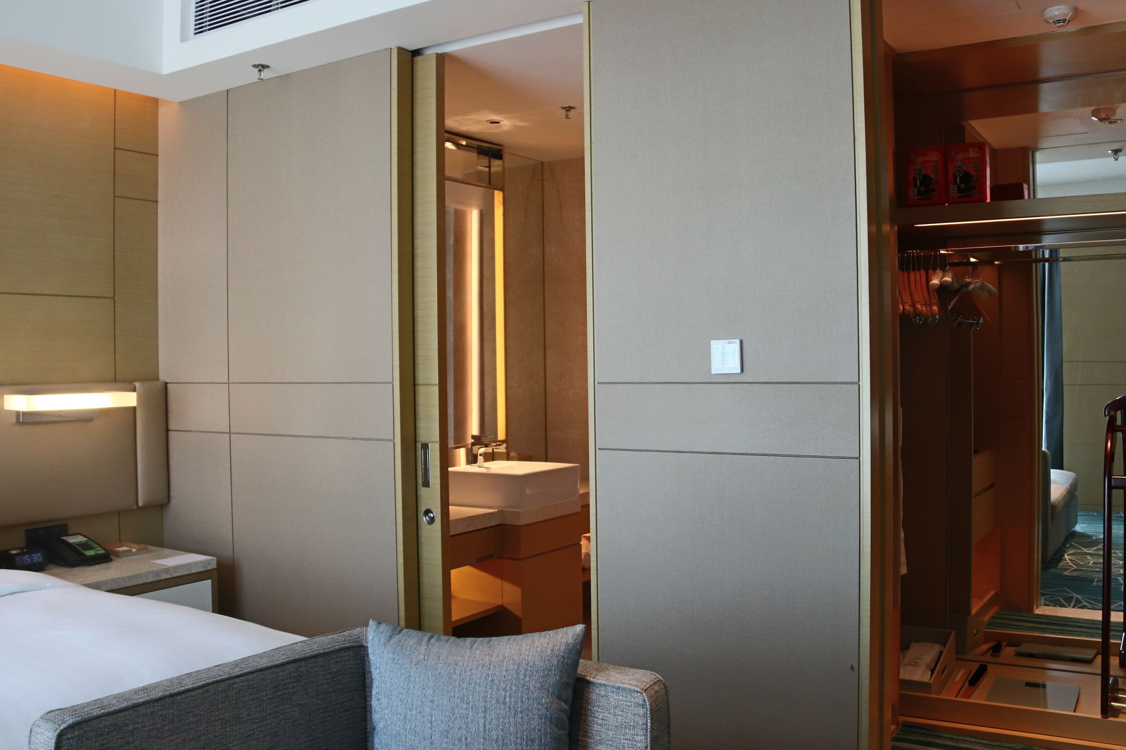 A hotel room with a bed, a bedside table with a phone, a mirror, a bathroom with a sink, and a shower area.