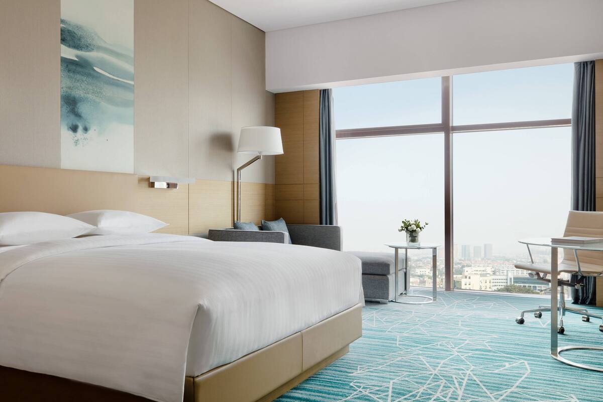 Quarto de hotel moderno com uma cama grande, uma escrivaninha, uma cadeira e vista para a cidade através de amplas janelas.