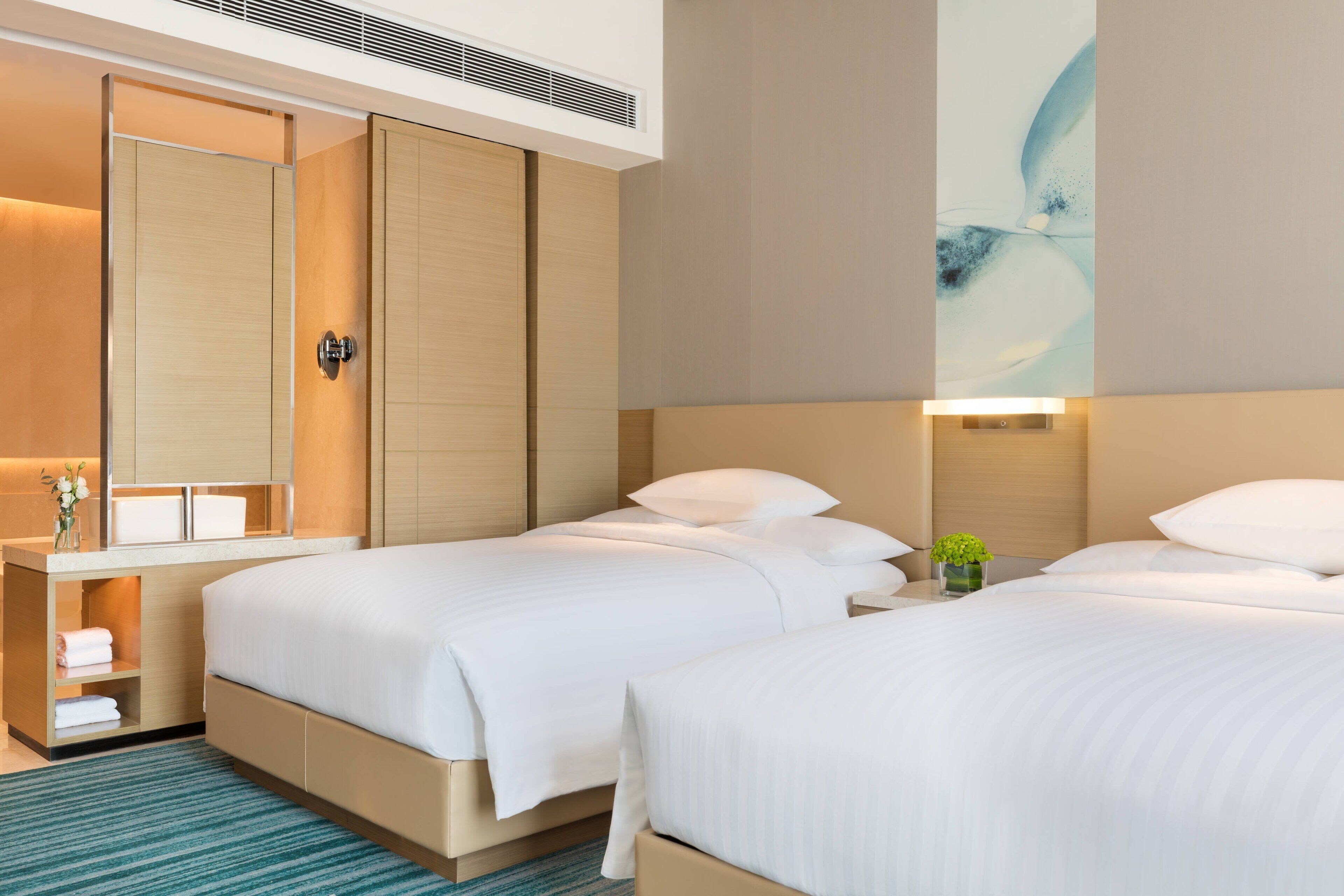 A hotel room with two beds, a wooden wardrobe, a bedside table with a lamp, and a wall-mounted artwork.