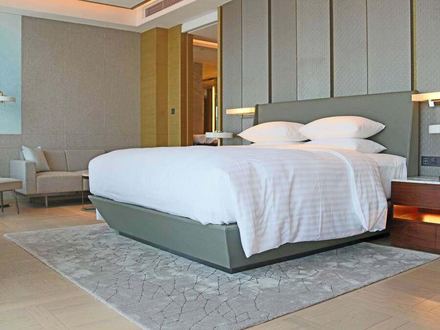 A modern hotel room with a large bed, a sofa, a desk, and a nightstand with a lamp.