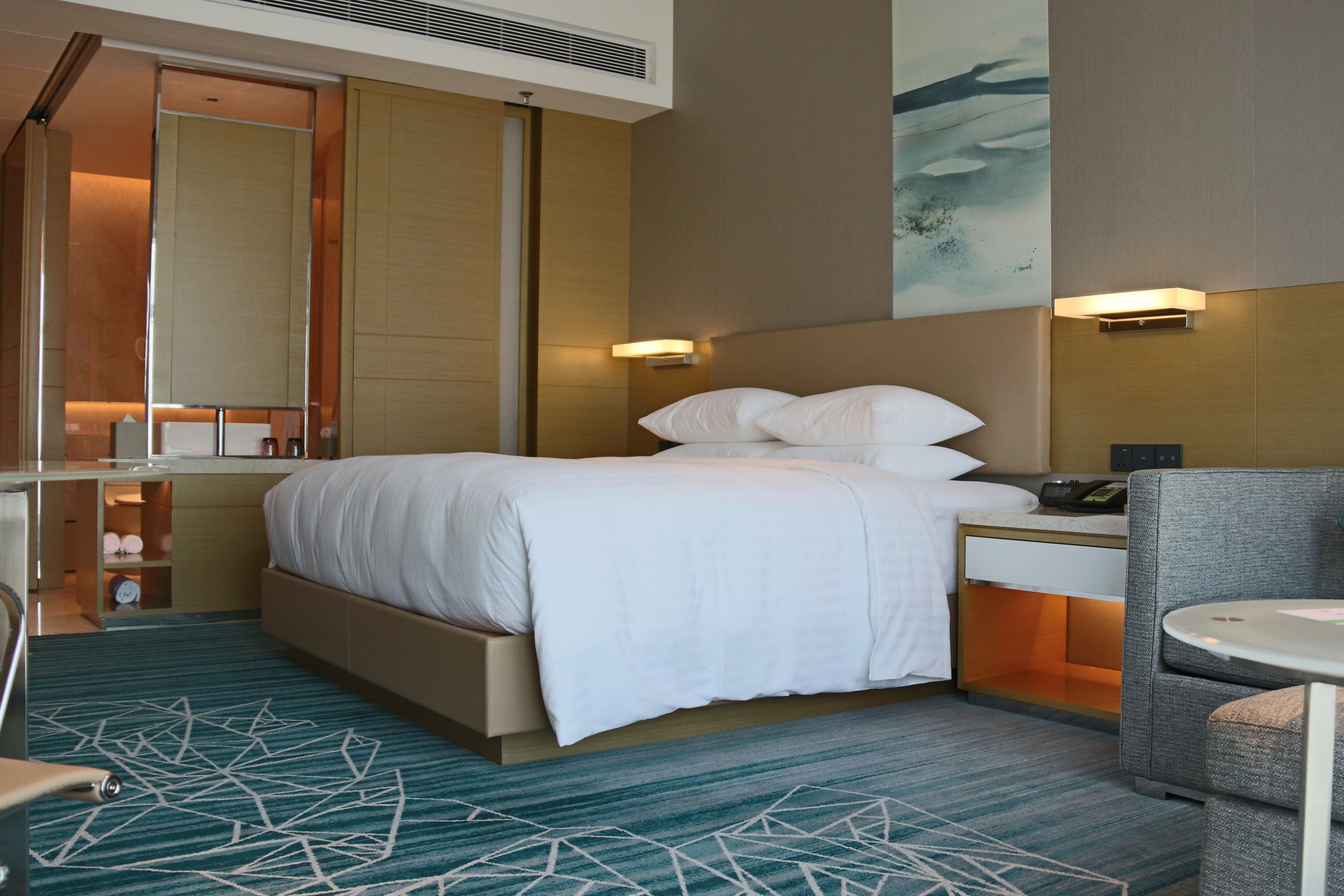 A hotel room with a bed, bedside table, chair, and a small table with a lamp.