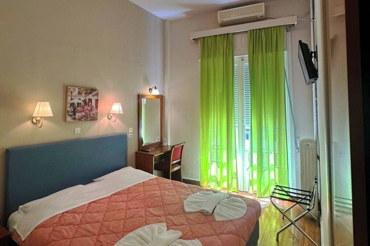 A hotel room with a bed, two bedside lamps, a desk, a chair, a TV, and a window with green curtains.