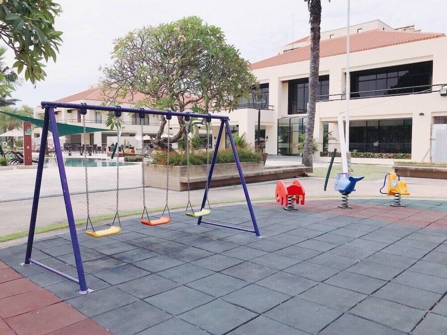 Ein Spielplatz mit Schaukeln und einer kleinen Rutsche, umgeben von einem befestigten Bereich und einem Gebäude mit rotem Dach.