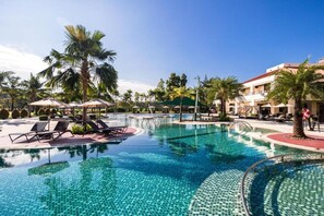 Ein Swimmingpool mit Liegestühlen und Sonnenschirmen, umgeben von Palmen und einem Gebäude mit Balkonen.