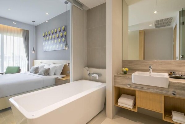 Una habitación de hotel moderna con una bañera grande, una cama doble, una mesita de noche y un espejo sobre el lavabo.