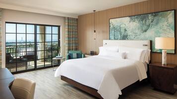 Una habitación de hotel moderna con una cama grande, vistas a través de ventanas de piso a techo, una zona de estar con sillas y una pared de acento de madera.
