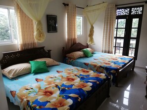 A room with two beds, each with floral bedding and a green pillow. There is a window with curtains, a framed picture on the wall, and a door with a decorative glass panel. - Abha Villa (Galle)
