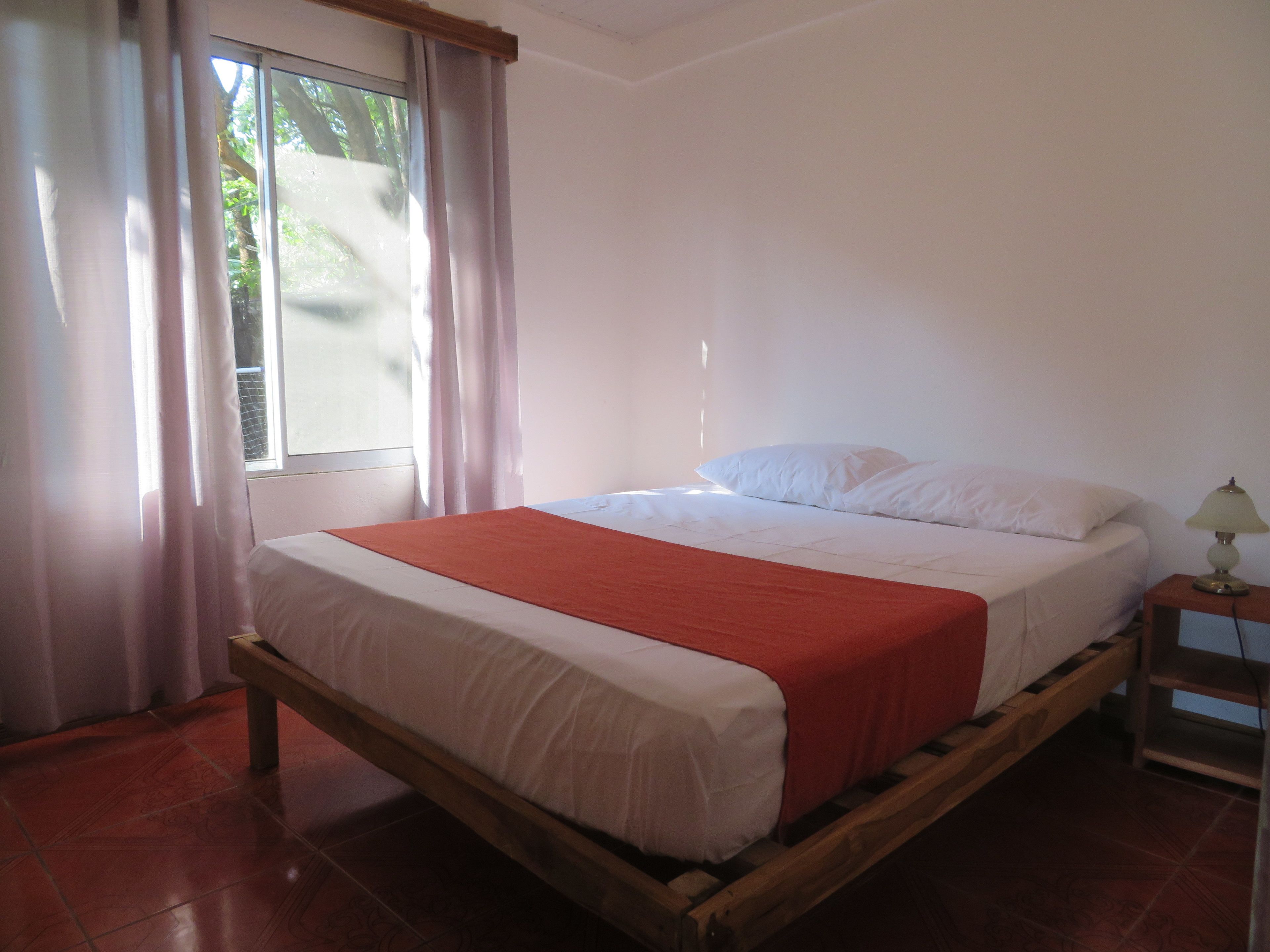 A single-bed room with a wooden frame, white bedding, a red blanket, a bedside table with a lamp, and a window with curtains.
