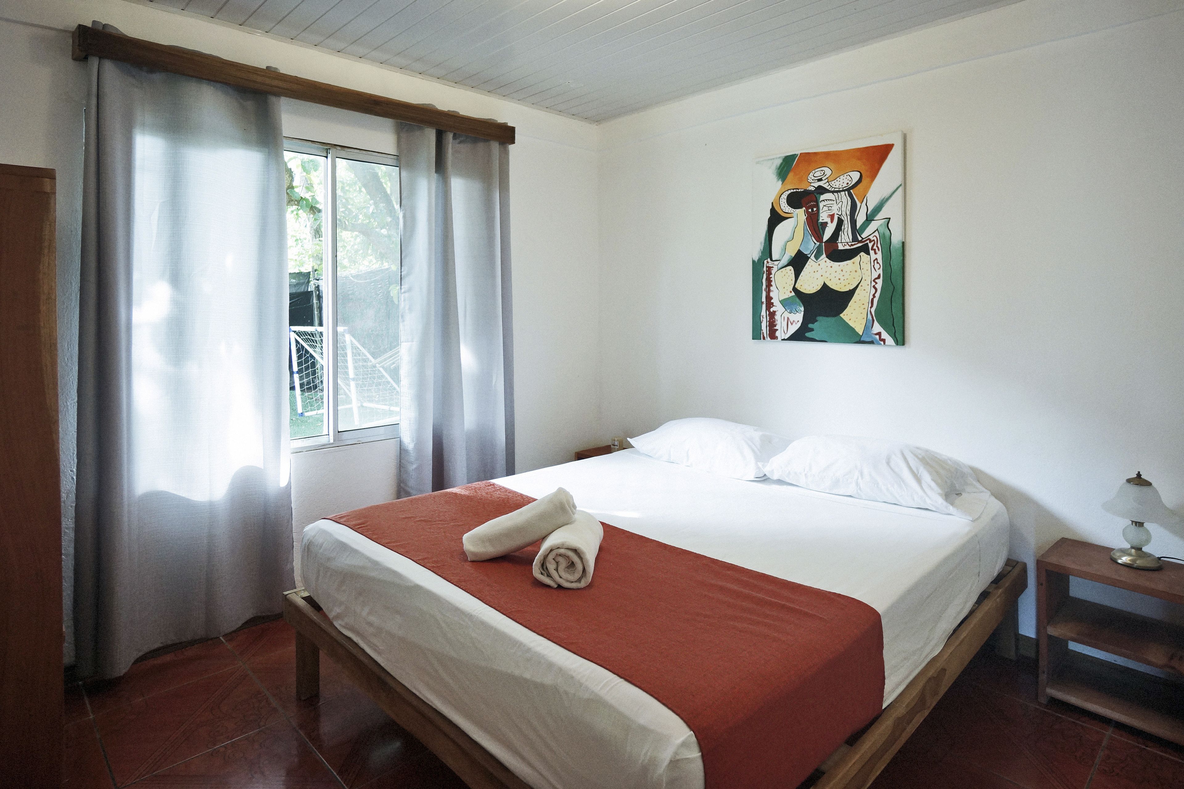 Una cama bien tendida con una colcha roja y dos toallas enrolladas, una ventana con cortinas, y un cuadro en la pared.