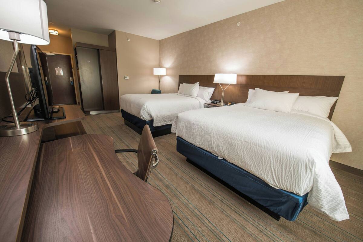 Ein Hotelzimmer mit zwei Betten, einem Schreibtisch und einem Stuhl. Auf dem Schreibtisch steht eine Schreibtischlampe und ein Computer.