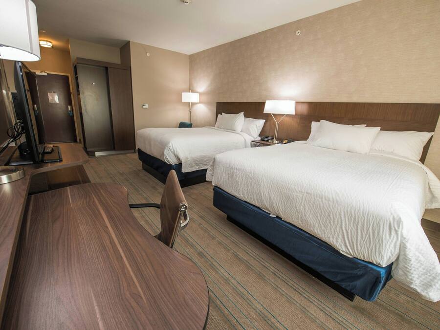 A hotel room with two beds, a desk, and a chair. There is a desk lamp and a computer on the desk.