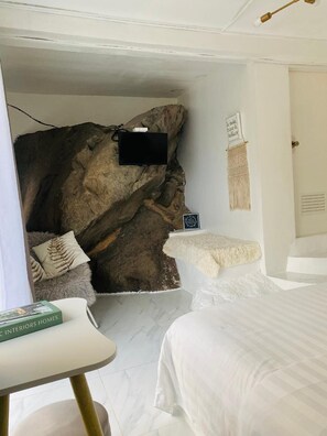 Un dormitorio con una gran pared rocosa, una cama, una mesita pequeña y un televisor fijado a la pared.