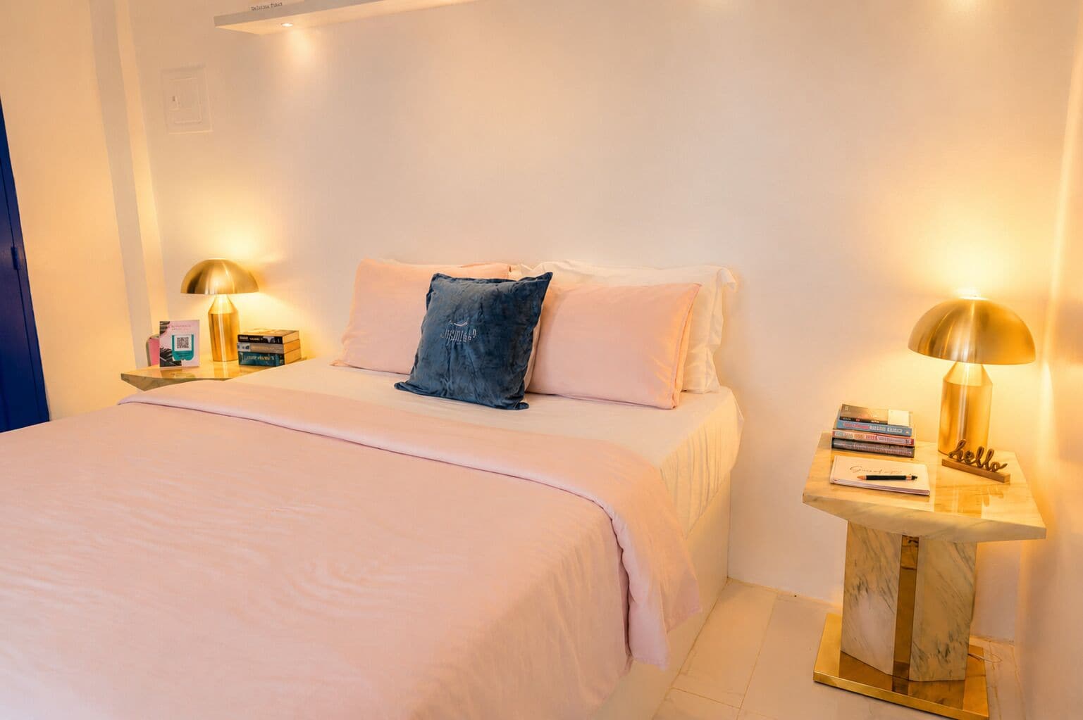 A hotel room with a bed, two bedside tables with lamps, a nightstand, and a small wooden side table with books and a notepad.