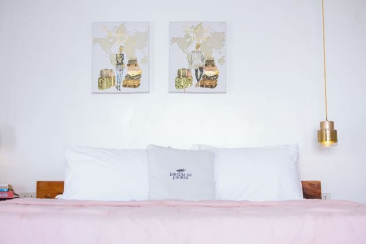 A bedroom with a bed, white pillows, a pink blanket, and two wall-mounted artworks depicting a person with luggage.