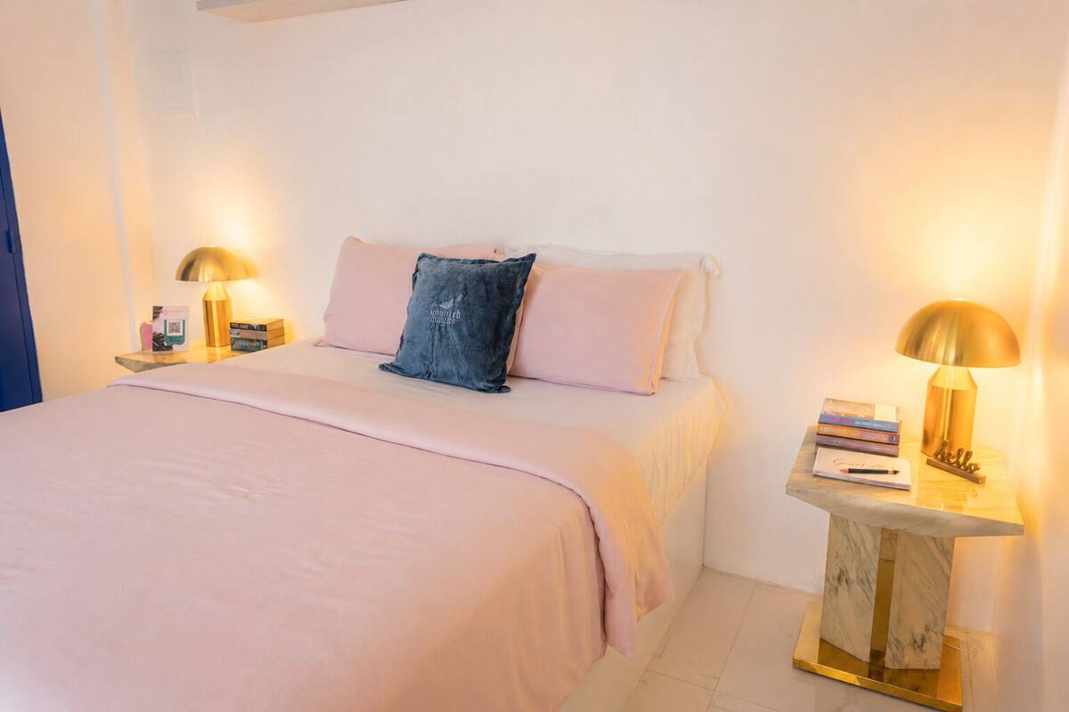 Un lit bien fait, avec une couvre-lit rose, deux oreillers et un coussin bleu. Il y a deux tables de chevet avec des lampes et des livres.