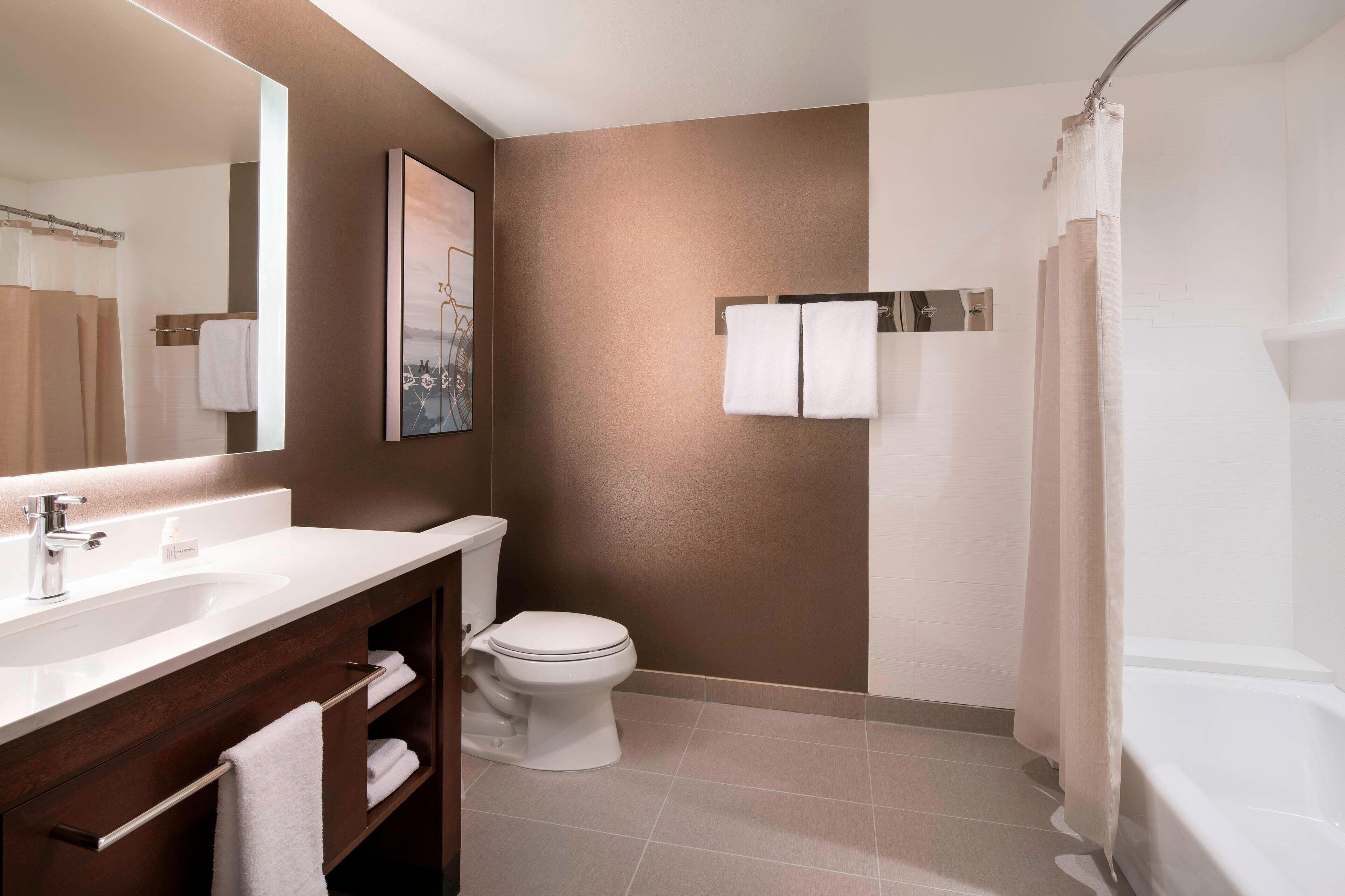 A modern bathroom with a white sink, toilet, and bathtub. There is a towel rack with two towels and a framed picture on the wall.