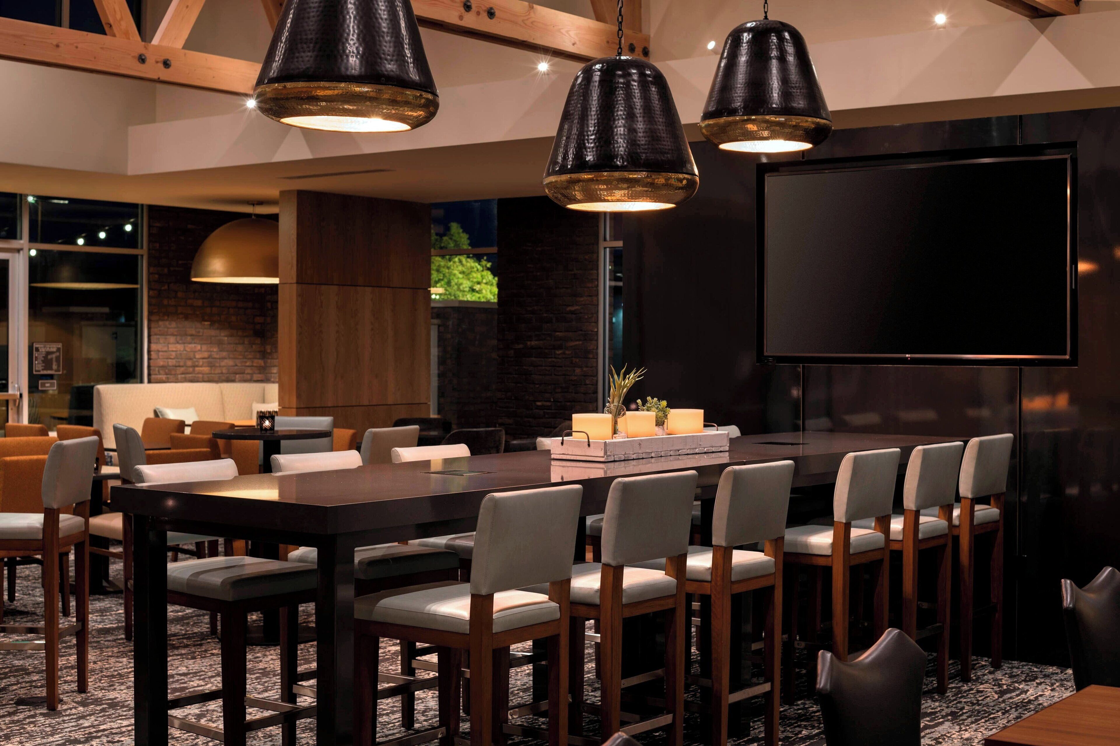 A modern conference room with a large table, multiple chairs, pendant lighting, a large screen, and a brick wall.