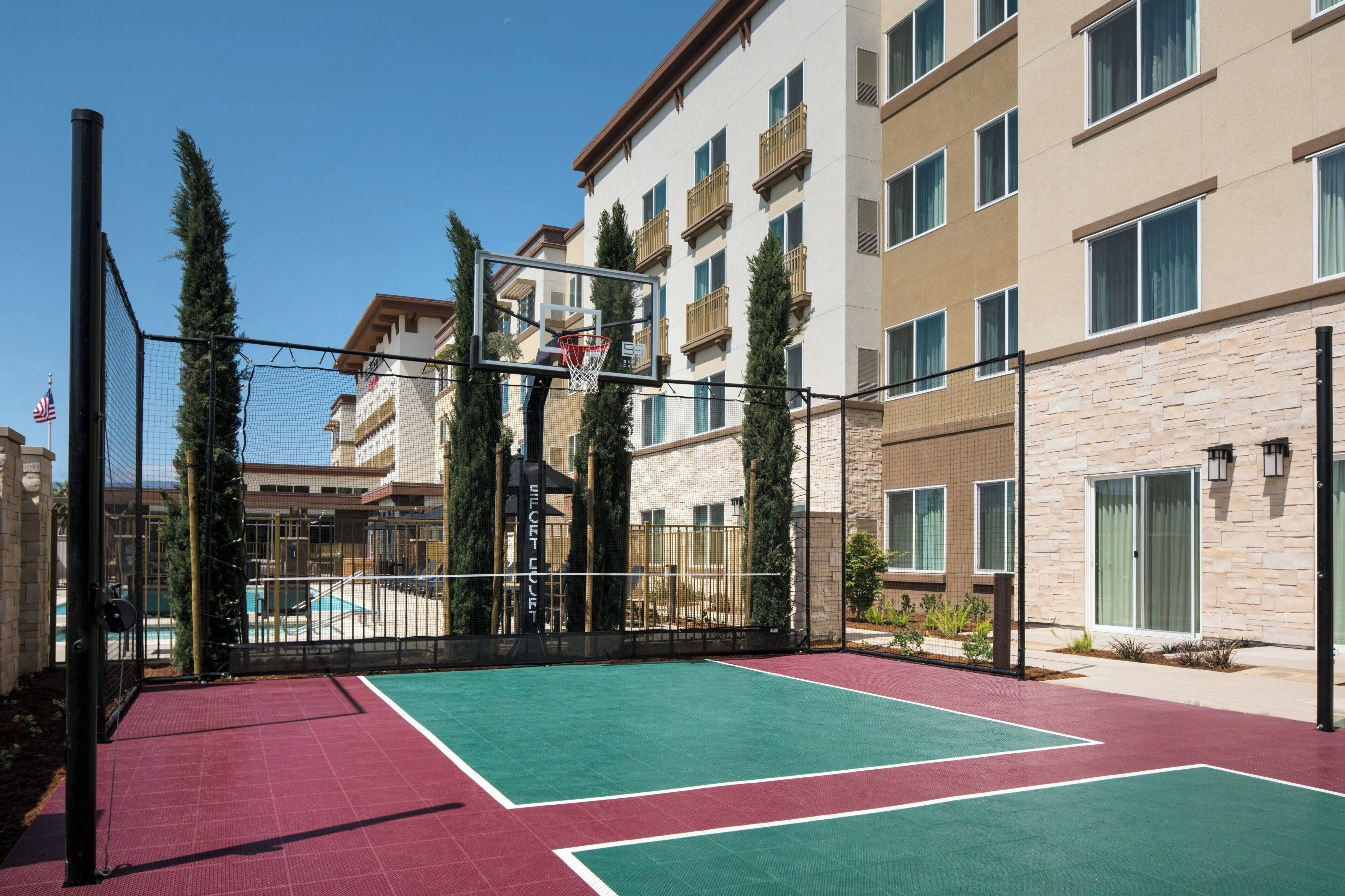 Un terrain de basket en plein air, situé à côté d’un immeuble d’habitation, avec une piscine en arrière-plan.