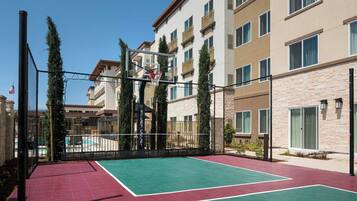 Un terrain de basket en plein air, situé à côté d’un immeuble d’habitation, avec une piscine en arrière-plan.