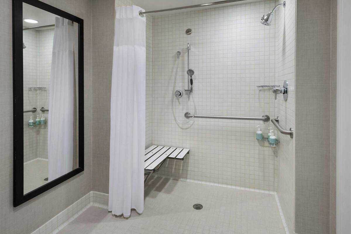 Un baño moderno con ducha a nivel, barras de apoyo, un banco y un espejo grande.