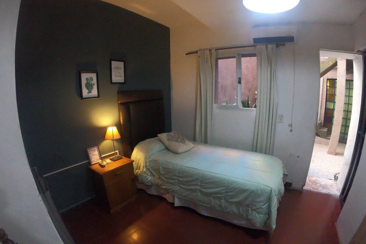 A bedroom with a bed, a nightstand with a lamp, a window with curtains, and a door leading to another room.