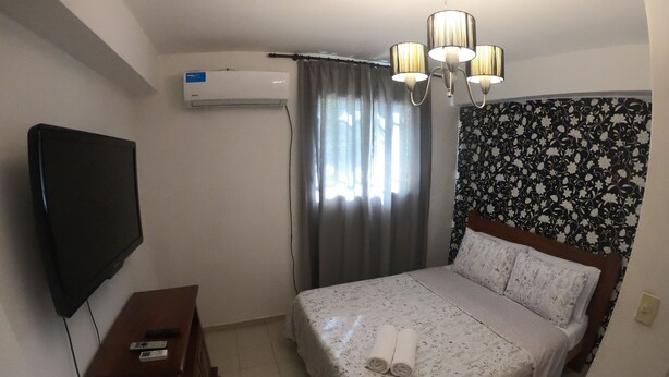 Hotellihuone, jossa on sänky, televisio, ilmastointilaite, puinen lipasto ja kuviollinen verho.