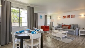A modern living room with a dining area, grey curtains, a red chair, a grey sofa, and artwork on the wall. - Dover Woods Apartments (St. Lawrence Gap)