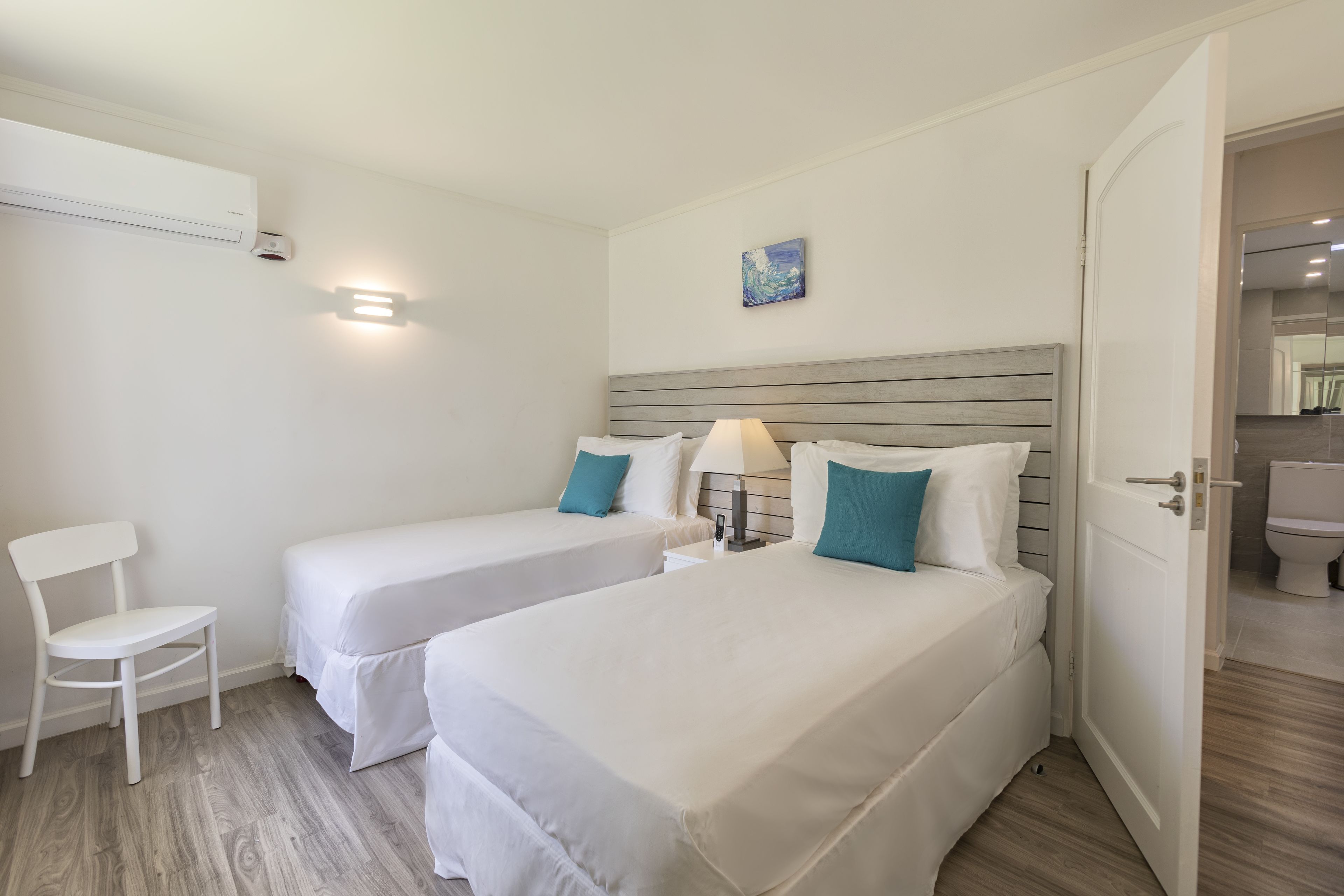 A hotel room with two single beds, a white chair, a bedside table with a lamp, and a painting on the wall.