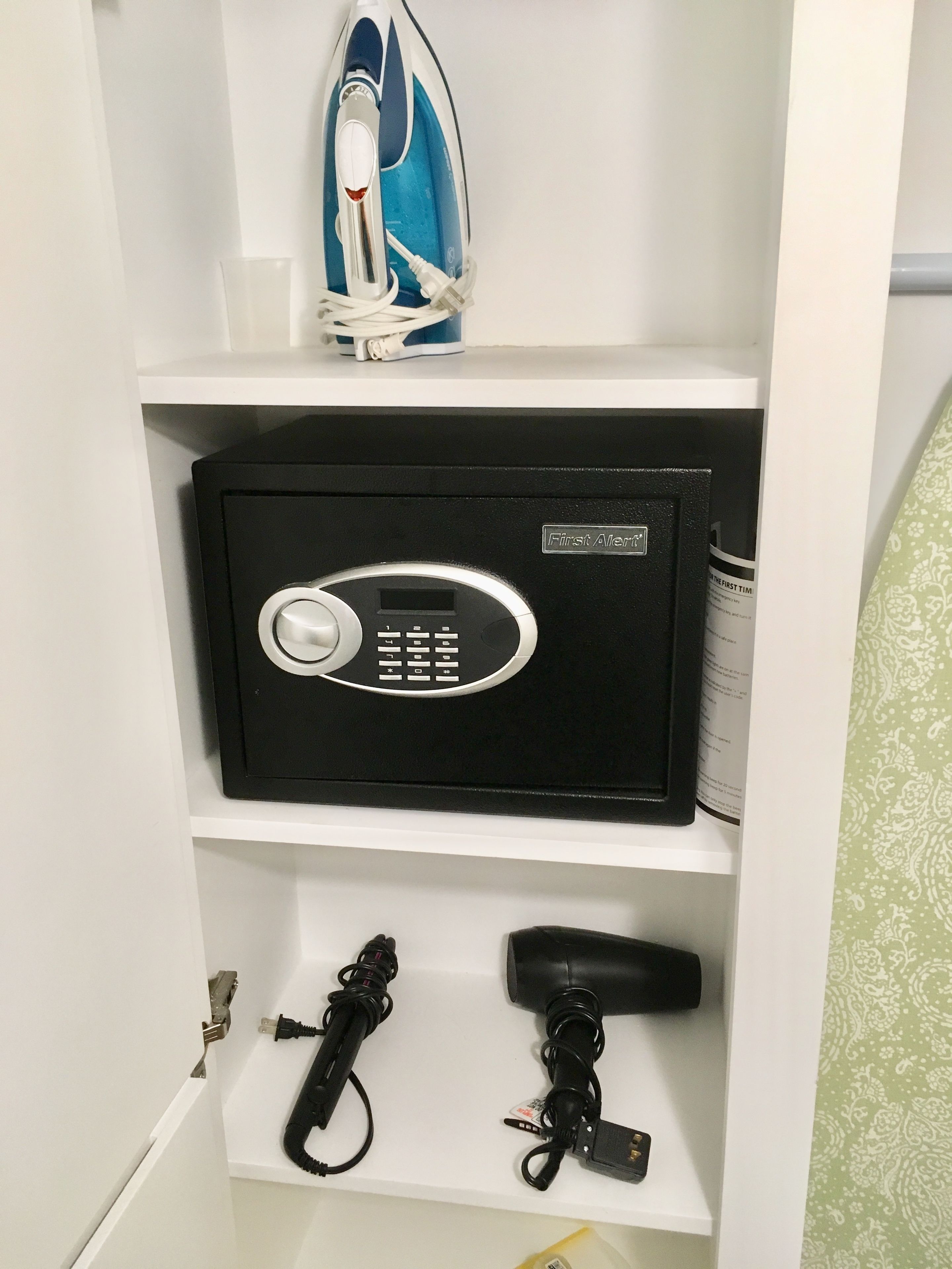 A white shelf with a black safe, a blue iron, and a hairdryer.