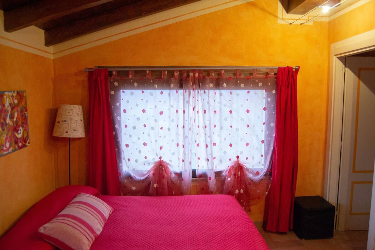 Um quarto com uma cama, janela com cortinas, abajur e um quadro na parede.