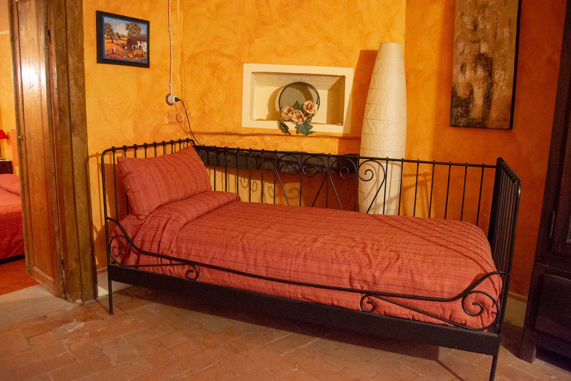Una cama individual con una colcha roja en una habitación con paredes naranjas, un cuadro enmarcado y un jarrón blanco con flores.
