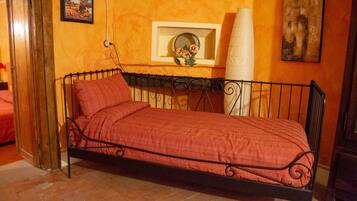Una cama individual con una colcha roja en una habitación con paredes naranjas, un cuadro enmarcado y un jarrón blanco con flores.