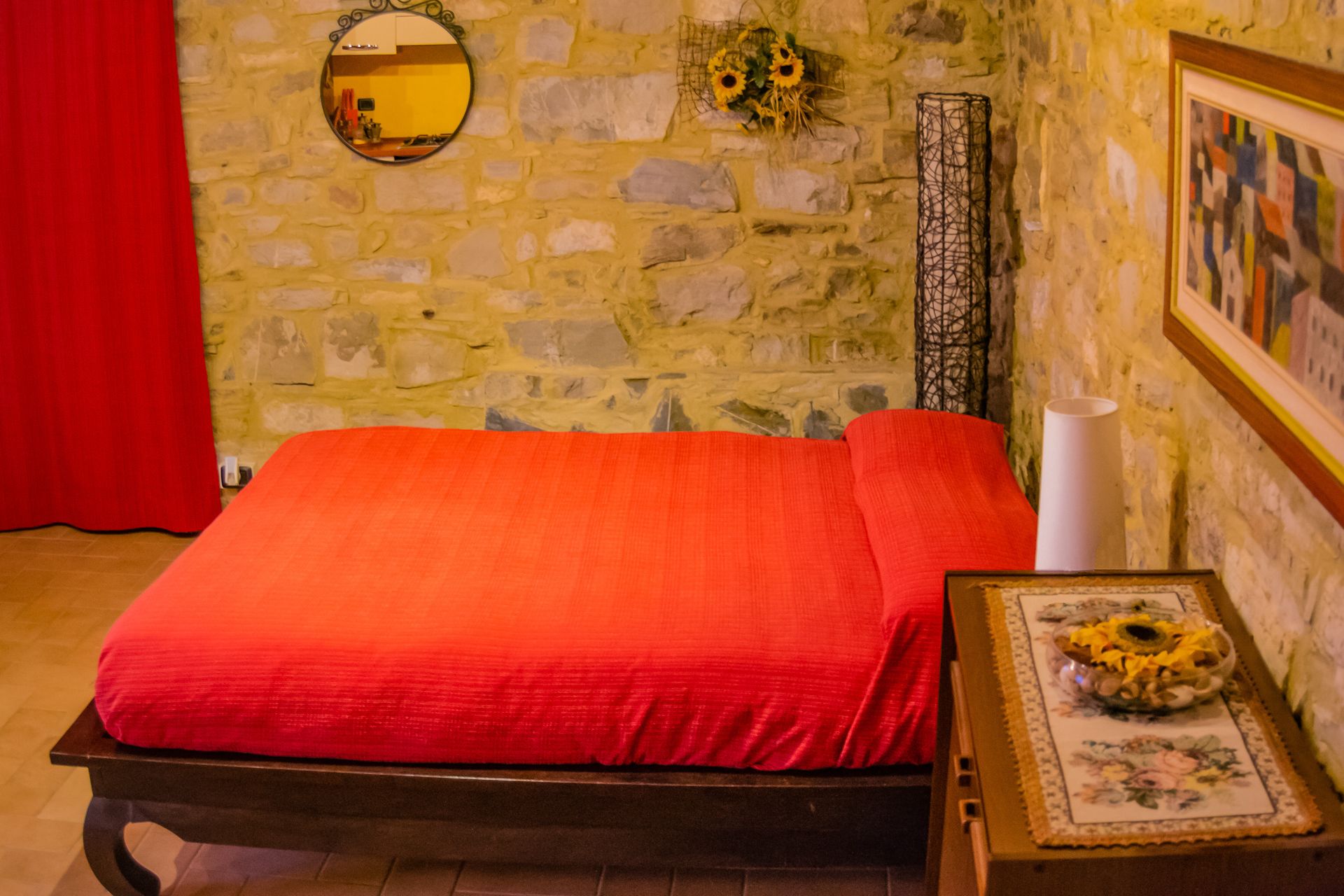 Une chambre avec un mur en pierre, un lit recouvert d’une couverture rouge, un miroir, un vase avec des tournesols et un tableau encadré.