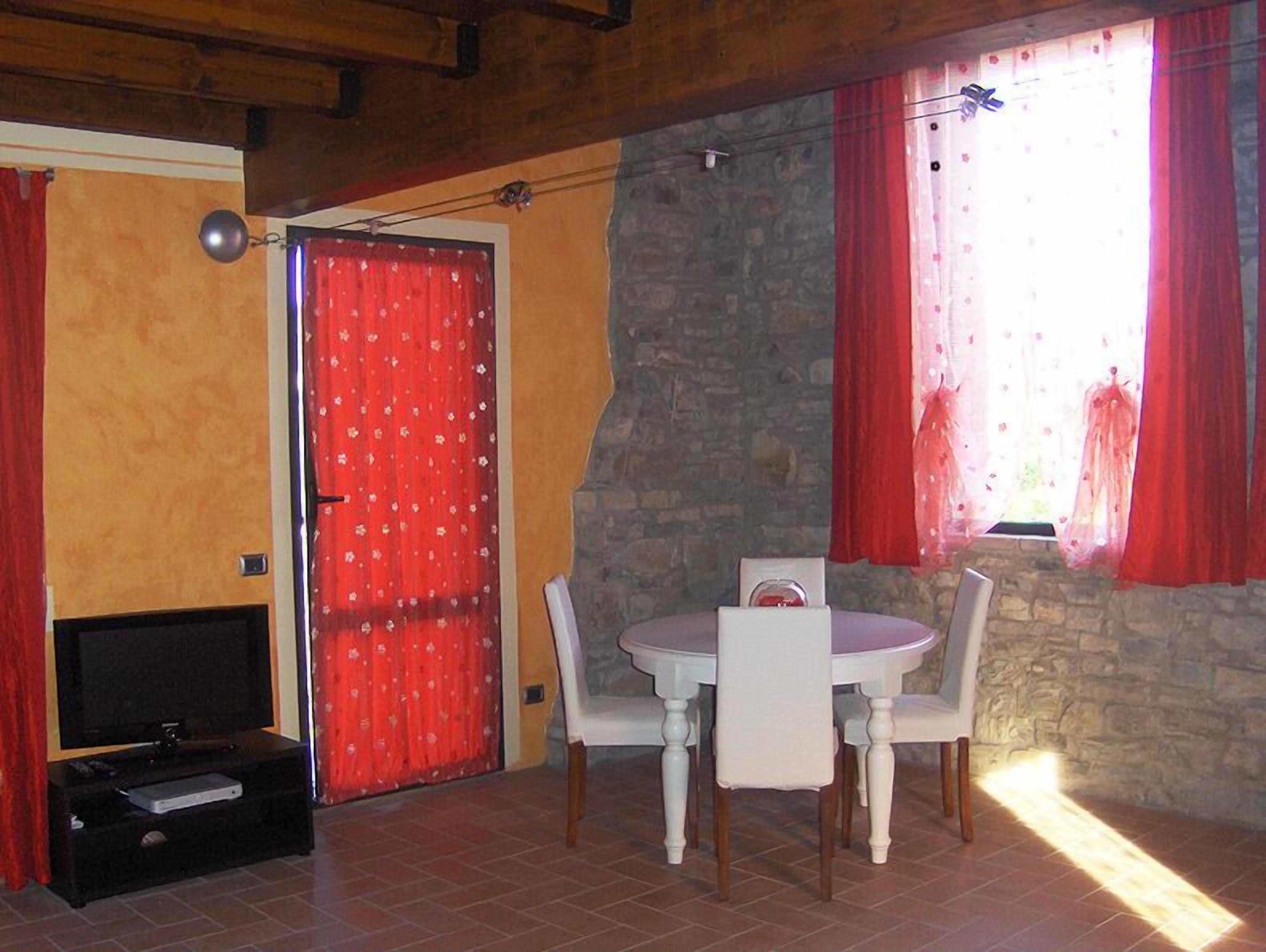 Une pièce avec un mur en pierre, une porte rouge, une table à manger avec des chaises et un téléviseur posé sur un meuble.