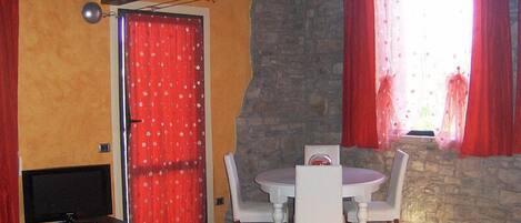 A room with a stone wall, a red door, a dining table with chairs, and a television on a stand.
