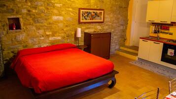 A room with a bed, a small cabinet, a painting on the wall, and a kitchenette area.