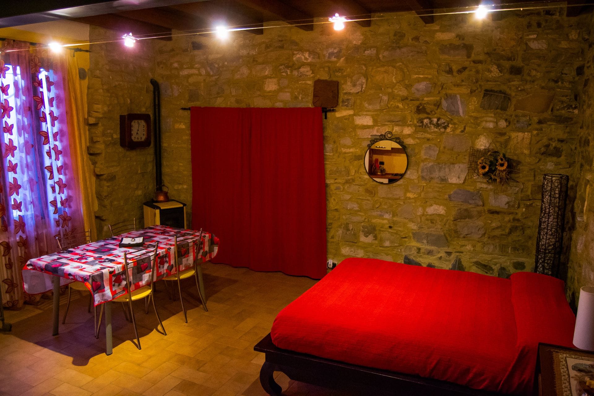 Une pièce avec un mur en pierre, un lit rouge, une table avec des chaises, une porte rouge et un miroir sur le mur.